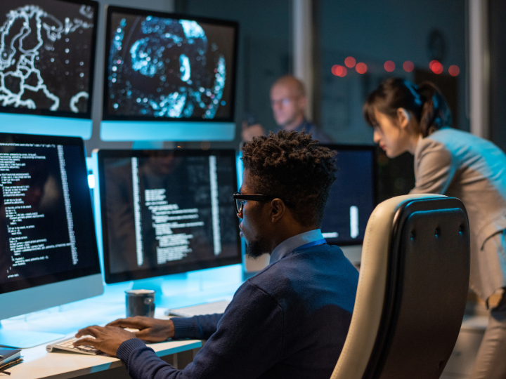 IT-ingenjör avkodar data medan han sitter framför datorskärmar och tittar på skärmen på en av dem.